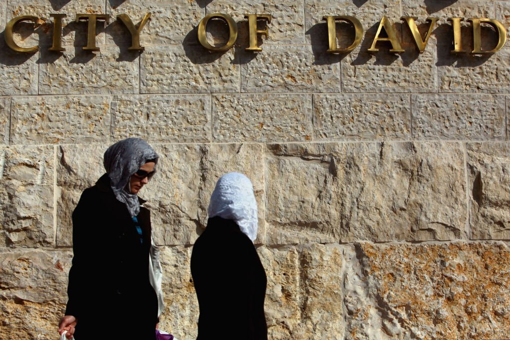 فلسطينيتان تمرّان أمام ما يُعرف بـ"مدينة داوود" وهي مركز استيطاني سياحيّ تسيطر عليه جمعية "إلعاد" الاستيطانية، يضم آثاراً تاريخية يعود بعضها للفترة الكنعانية، وفيها عين سلوان وغيرها من الآثار. (Getty Images).